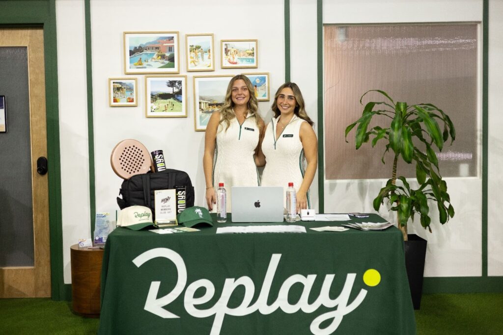 Two people smiling and standing behind a Replay Club-branded table during a sports event. 