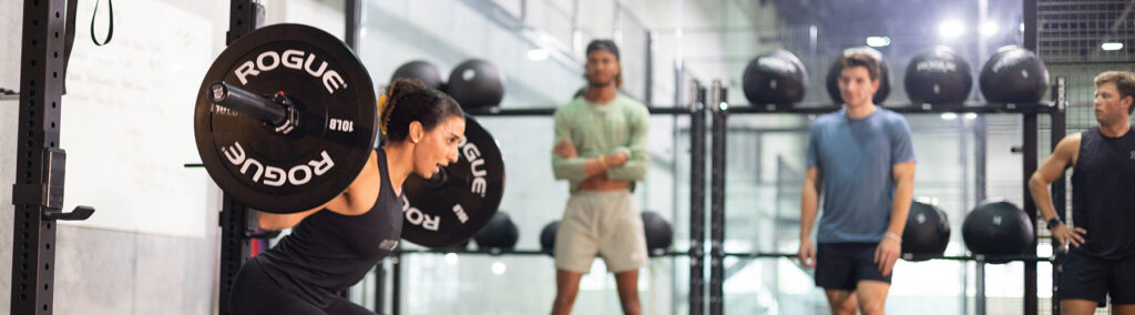 A woman performs a barbell back squat at a Rogue-equipped gym while three observers watch in the background, illustrating the supportive community environment at Replay Club.